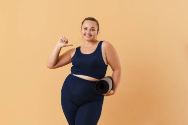 Bej arka planda izole edilmiş spor minderiyle poz verirken kendini gösteren genç bir kadın.