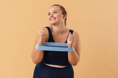 Spor takım elbiseli genç bir kadın, bej arka planda izole edilmiş genişletici gerilimle egzersiz yapıyor.