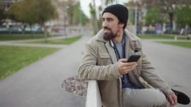 Şapka takan yakışıklı sakallı adam dışarıda telefonla yazı yazıyor.