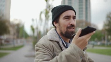 Şapkalı, gülen sakallı adam dışarıda telefonla sesli mesaj kaydediyor.