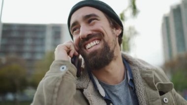 Şapkalı neşeli sakallı adam dışarıda telefonla konuşuyor.