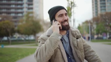 Şapkalı, gülen sakallı adam dışarıda telefonla konuşuyor.