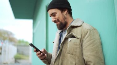 Şapka takan mutlu sakallı adam dışarıda telefonla yazı yazıyor.