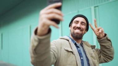 Şapkalı, gülümseyen sakallı adam dışarıdaki telefonlara barış işareti yapıyor.