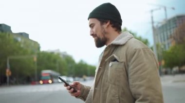 Ciddi sakallı bir adam şehirde yürürken ve cep telefonunu tutarken.
