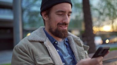 Şehirde telefonla mesajlaşan, şapkalı, gülen adam.