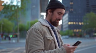 Şapkalı mutlu bir adam cep telefonunda daktilo ediyor ve şehirdeki kameraya bakıyor.