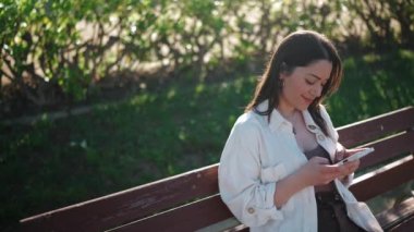 Gülümseyen esmer kadın bankta otururken cep telefonunda yazıyor.