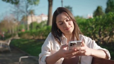 Tebessüm eden esmer kadın bankta otururken telefonla konuşuyor.