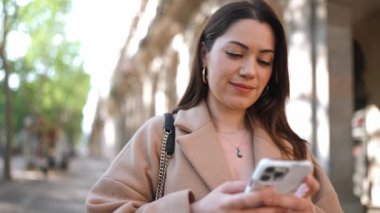 Gülümseyen esmer kadın telefonda yazıyor ve caddedeki kameraya bakıyor.