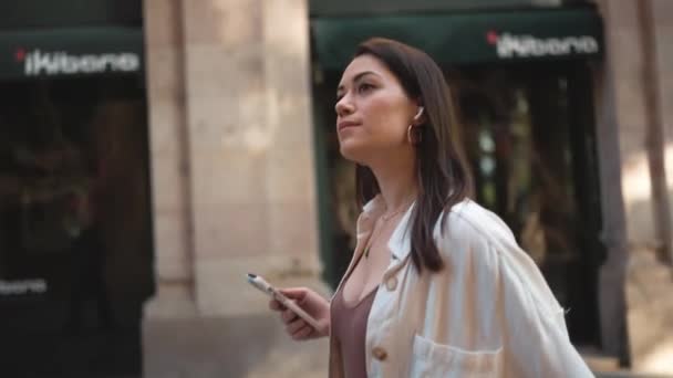 漂亮的黑发女人戴着耳机在街上走路和拿着手机 — 图库视频影像