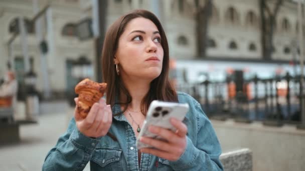 漂亮的黑发女游客吃着面包 坐在长椅上看手机 — 图库视频影像