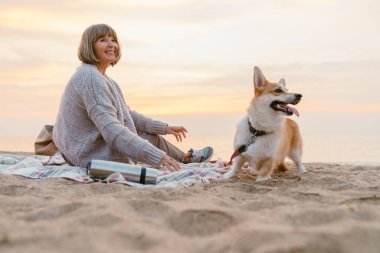 Yaşlı kadın köpeğiyle kumsalda dinlenirken gülümsüyor.
