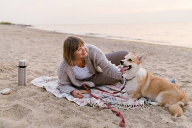 Yaşlı kadın köpeğiyle kumsalda dinlenirken gülümsüyor.