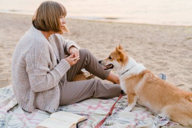 Yaşlı kadın, köpeğiyle kumsalda dinlenirken kitap okuyor.