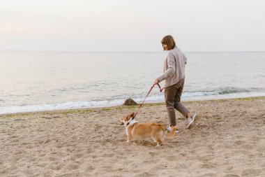 Yaşlı kadın kumsalda köpeğini gezdirirken gülümsüyor.