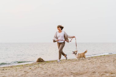 Yaşlı kadın kumsalda köpeğini gezdirirken gülümsüyor.