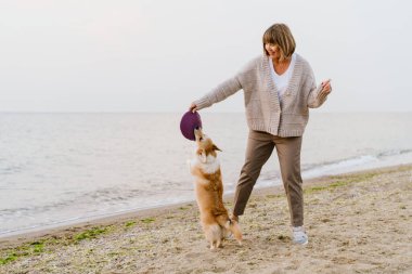 Yaşlı kadın kumsalda köpeğiyle oynarken gülümsüyor.