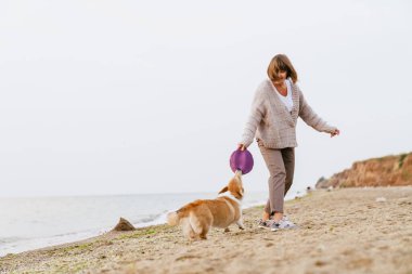 Yaşlı kadın kumsalda köpeğiyle oynarken gülümsüyor.