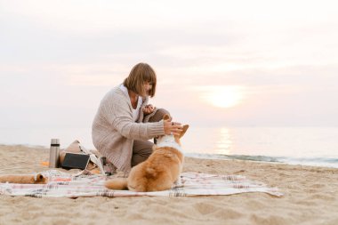 Yaşlı kadın köpeğiyle kumsalda dinlenirken gülümsüyor.