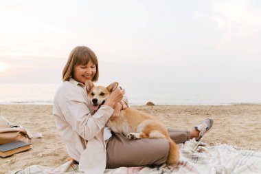 Yaşlı kadın köpeğiyle kumsalda dinlenirken gülümsüyor.