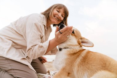 Yaşlı kadın köpeğiyle kumsalda dinlenirken gülümsüyor.