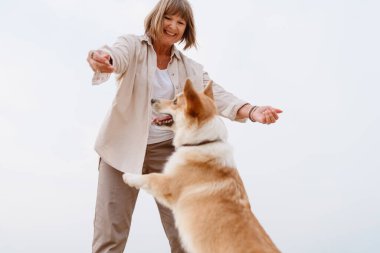 Yaşlı kadın kumsalda dinlenirken köpeğiyle gülüp oynuyor.