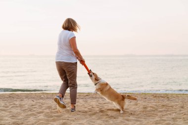 Yaşlı Avrupalı kadın kumsalda köpeğiyle oynuyor.
