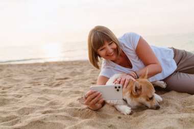 Son sınıf öğrencisi, kumsalda köpeğiyle dinlenirken cep telefonuyla fotoğraf çekiyor.