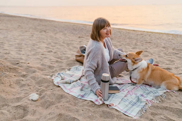Yaşlı kadın köpeğiyle kumsalda dinlenirken gülümsüyor.