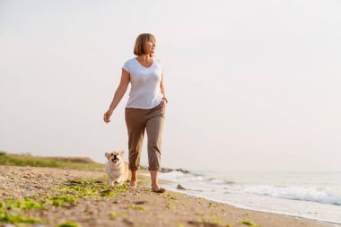 Yaşlı kadın kumsalda köpeğini gezdirirken gülümsüyor.
