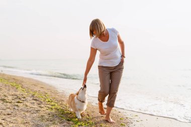Yaşlı kadın kumsalda köpeğini gezdirirken gülümsüyor.