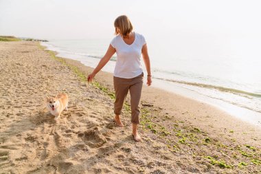 Yaşlı kadın kumsalda köpeğini gezdirirken gülümsüyor.
