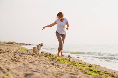 Yaşlı kadın kumsalda köpeğini gezdirirken gülümsüyor.