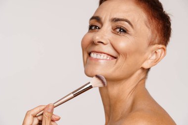 Beyaz arka planda izole edilmiş toz fırçası kullanan kısa saçlı yaşlı üstsüz kadın.