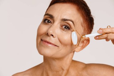 Beyaz arka planda izole edilmiş yeşim taşlı yüzle masaj yaparken kıdemli üstsüz kadın gülümsüyor.