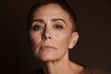 Kızıl saçlı, kısa saçlı, kahverengi duvarda izole edilmiş kameraya bakan yaşlı bir kadın.