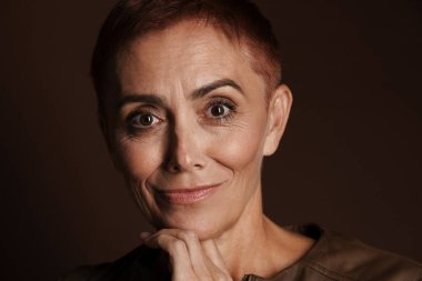 Kıdemli kızıl saçlı, kısa saçlı, gülümseyen ve kahverengi duvardan izole edilmiş kameraya bakan kadın.