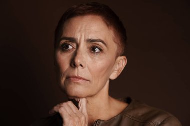 Kızıl saçlı, kısa saçlı yaşlı bir kadın kahverengi duvarın üzerinde tek başına poz veriyor ve bakıyor.