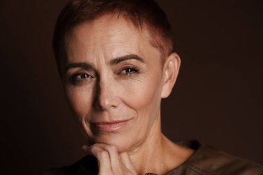 Kızıl saçlı, kısa saçlı yaşlı bir kadın kahverengi duvarın üzerinde tek başına poz veriyor ve bakıyor.