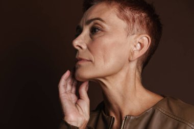 Kızıl saçlı, kısa saçlı yaşlı bir kadın kahverengi duvarın üzerinde tek başına poz veriyor ve bakıyor.