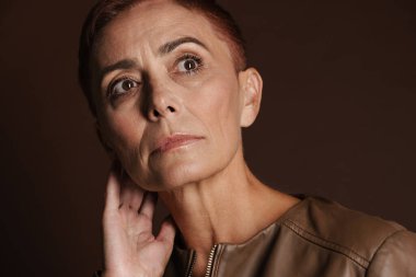 Kızıl saçlı, kısa saçlı yaşlı bir kadın kahverengi duvarın üzerinde tek başına poz veriyor ve bakıyor.