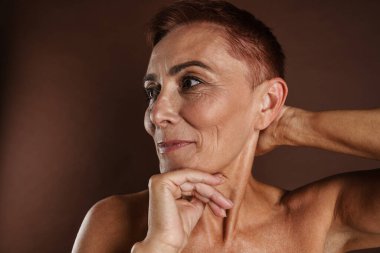 Kahverengi duvarın üzerinde yalnız poz verirken gülümseyen son sınıf üstsüz kadın.