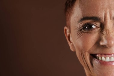 Kahverengi duvarın üzerinde yalnız poz verirken gülümseyen son sınıf üstsüz kadın.