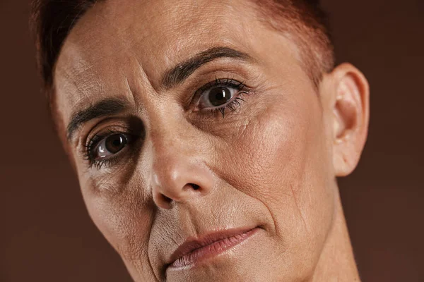 Kızıl saçlı, kısa saçlı, kahverengi duvarda izole edilmiş kameraya bakan yaşlı bir kadın.