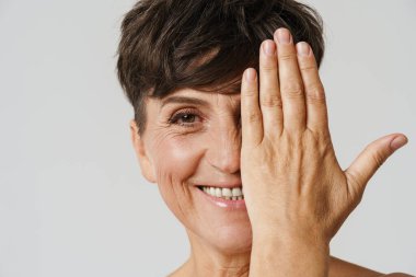 Yetişkin yarı çıplak bir kadın, gülümserken beyaz arka planda izole edilmiş gözünü kapatıyor.