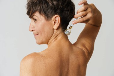 Olgun yarı çıplak bir kadın beyaz arka planda izole edilmiş yeşim taşıyla boynuna masaj yapıyor.