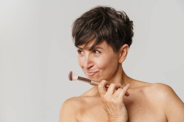 Beyaz arka planda toz fırçasıyla poz verirken gülümseyen olgun yarı çıplak kadın.