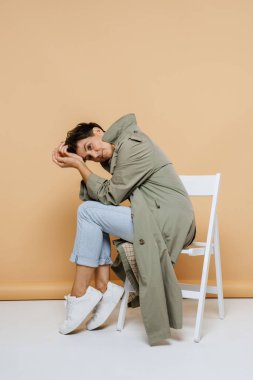 Olgun Avrupalı kadın bej arka planda izole edilmiş bir sandalyede otururken kameraya bakıyor