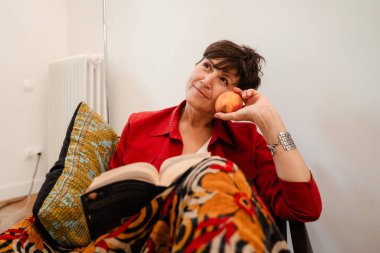 Olgun esmer kadın evde kanepede otururken kitap okuyor.